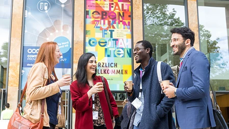 Vier lächelnde Menschen in Business-Kleidung stehen beim 103. Deutschen Röntgenkongress vor dem RheinMain CongressCenter (RMCC) in Wiesbaden. Sie haben Kaffeebecher in der Hand und unterhalten sich.