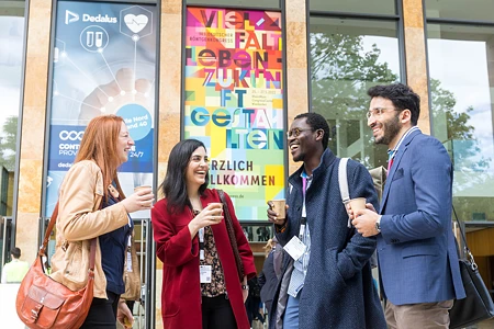 Vier lächelnde Menschen in Business-Kleidung stehen beim 103. Deutschen Röntgenkongress vor dem RheinMain CongressCenter (RMCC) in Wiesbaden. Sie haben Kaffeebecher in der Hand und unterhalten sich.