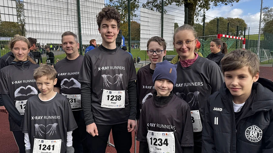 Eine Gruppe von Jugendlichen in schwarzen Laufshirts mit der Aufschrift 'X-Ray Runners' und Startnummern steht auf einem Sportplatz.